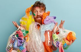 Photo of surprised red haired man has thick beard, overloaded with much garbage, collects plastic, looks with frustrated expression, raises hands, isolated oer blue background. Ecology concept